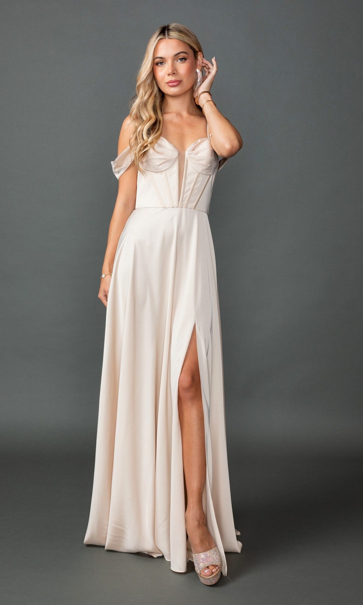 A woman wears the Dancing Queen 4544 long prom dress, a light beige, off-the-shoulder gown with a thigh-high slit and lace-up back, standing against a plain gray background.